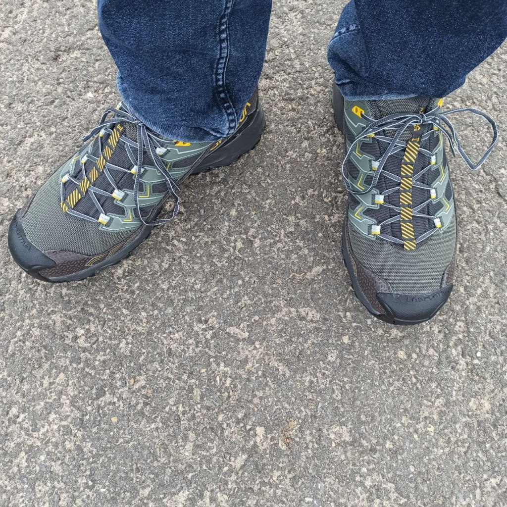 Die gleichen grün-grauen Schuhe, wie sie an meinen Füßen sitzen. Das Bild ist von oben gemacht. Man sieht den grauen Asphalt des Weges und die Hosenbeine meiner dunkelblauen Jeans.